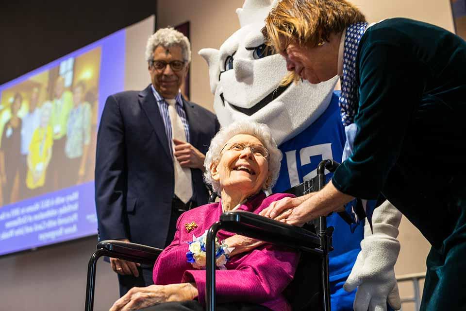 Two people stand and one person sits on a stage near the Billiken.