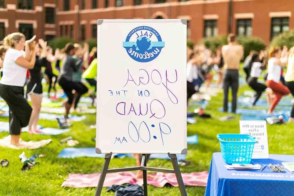 People practicing yoga in background with sign reading "Yoga on the Quad, 9 a.m." in the foreground.