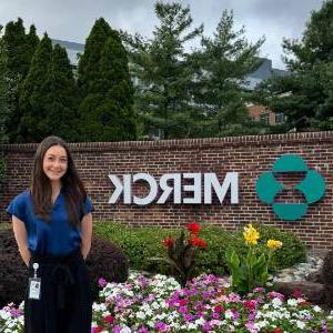 Lauren Favell stands in front of a flower bed and a large sign that says Merck.